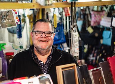 A male stallholder wearing glasses smiles at the camera from behind the table of wallets and purses for sale