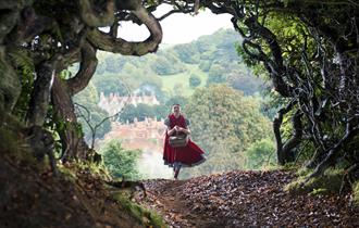 A lady in a red dress carries a basket through the woods with a the sunny countryside behind her on the horizon.