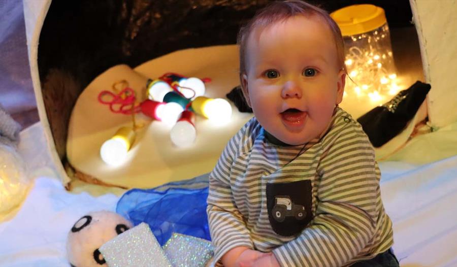 A happy baby faces the camera, sitting on the floor and surrounded by various items including jars of lights.