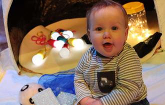 A happy baby faces the camera, sitting on the floor and surrounded by various items including jars of lights.