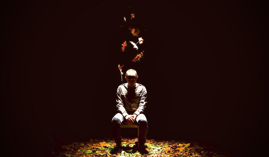Image credit Drew Forsyth.  A man sit on a chair on the stage surrounded in darkness with the spotlight on him.  Autumn leaves fall from above and are
