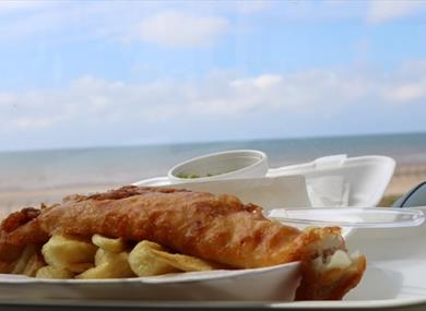 A plate of fish and chips against the backdrop of Blackpool beach on a sunny day.