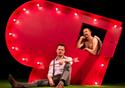 A performer sits on stage next to a large red heart, holding cupid arrow.  Cupid looks on through a small window in the heart.  Credit Zoe Manders