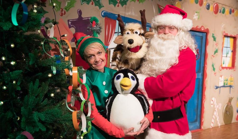 Santa poses in his workshop with an elf, a penguin and a reindeer.