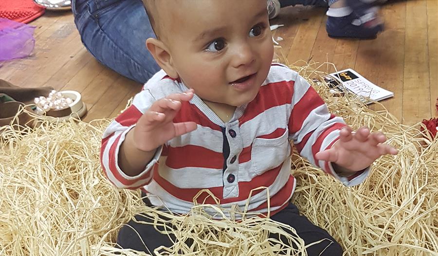 A happy toddler is sat amongst straw like material, reaching out to touch it.