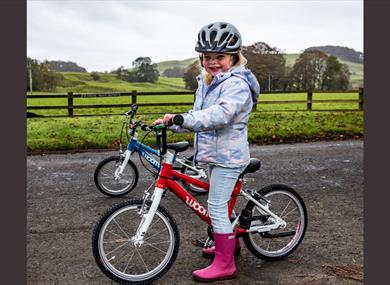 A small child in pink wellies, rides a bike with the countryside in the background.