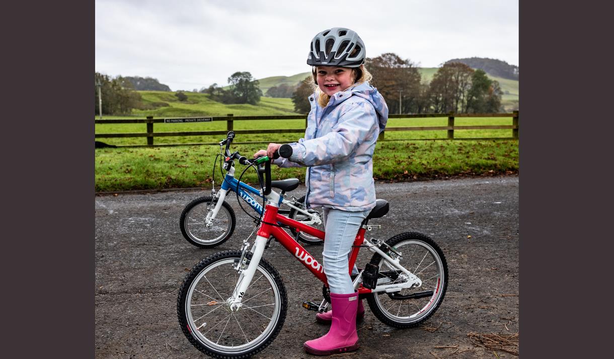 A small child in pink wellies, rides a bike with the countryside in the background.