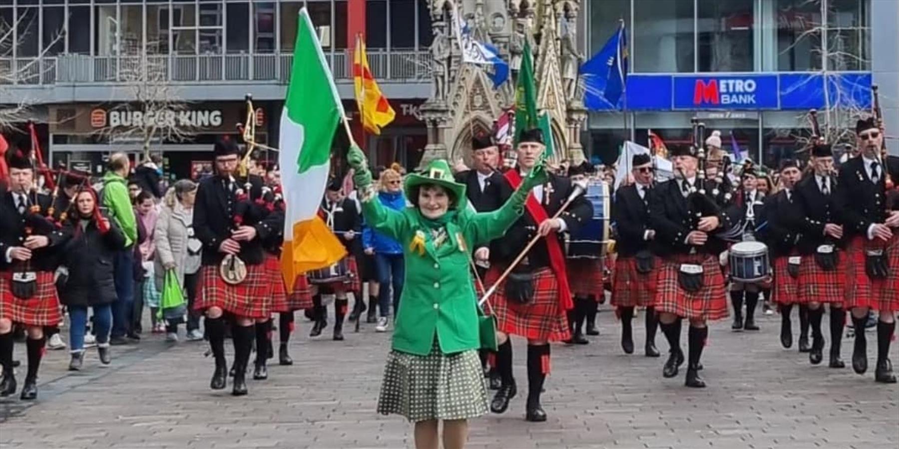 A lady with Irish flag and green suit in front of a pipe and drums band
