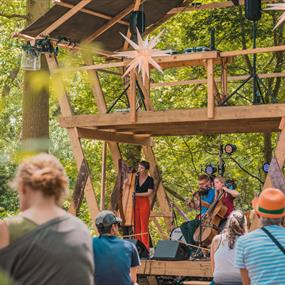 A musician with red trousers stands on the Eyrie Stage at Timber Festival surrounded by trees and an audience