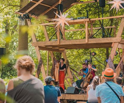 A musician with red trousers stands on the Eyrie Stage at Timber Festival surrounded by trees and an audience 