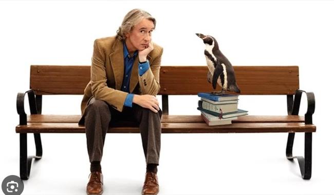 Actor Steve Coogan sitting on a bench with a penguin on a pile of books