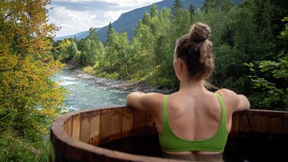 Person sitting in a wooden outdoor hot tub beside a river, with forest and mountains in the background.