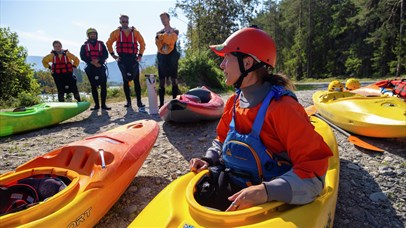 Group of kayaks gathered on the riverbank, with participants wearing helmets, life jackets, and paddling gear.