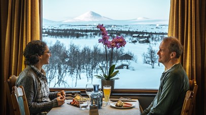 A couple eat a meal in front of a window with a view of snowy mountains