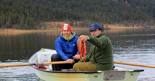 Two men in a rowboat. One steers the boat and the other shows the catch.