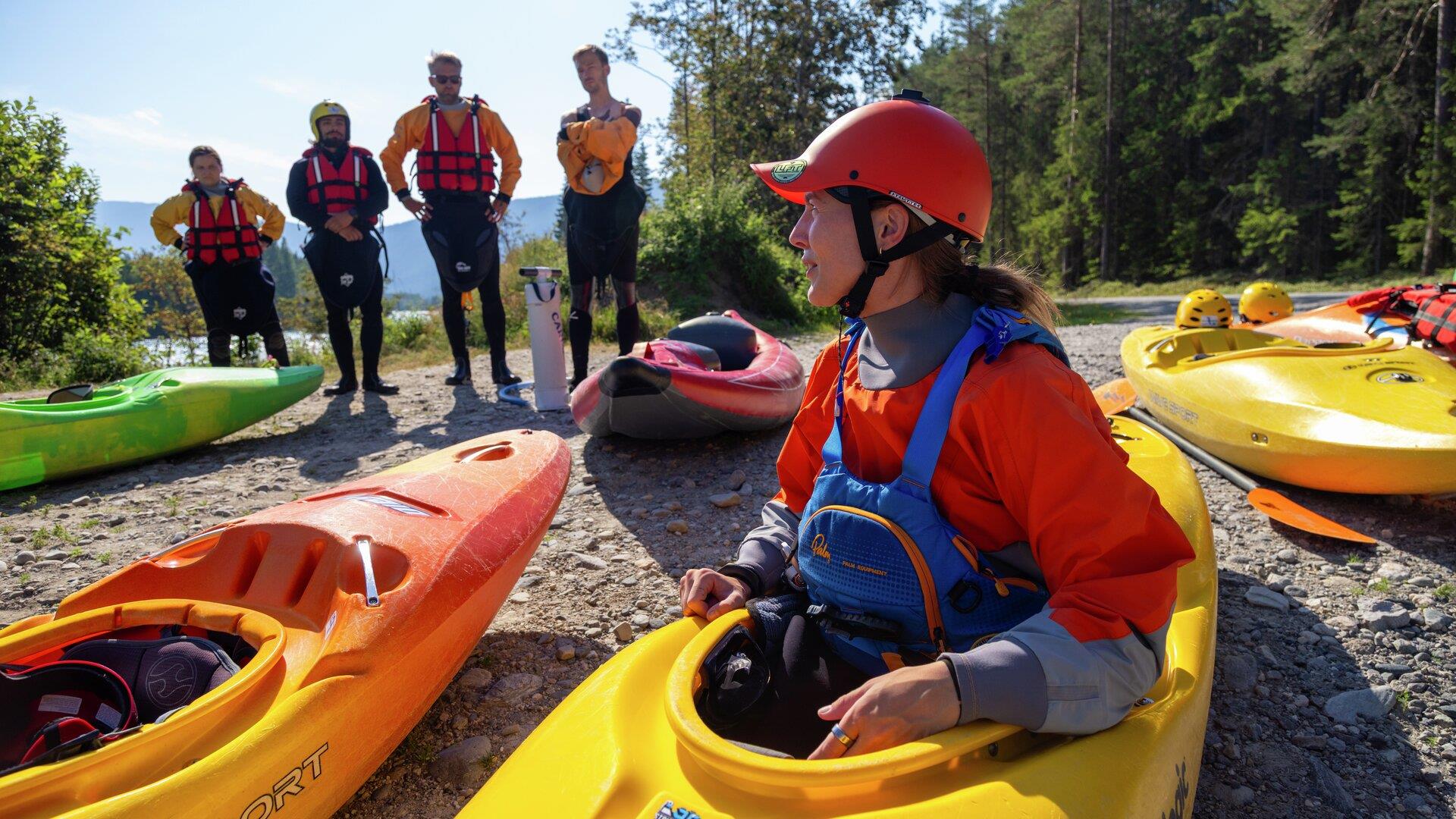 Gruppe med kajakker samlet på elvebredden, med deltakere iført hjelm, redningsvest og padleutstyr.