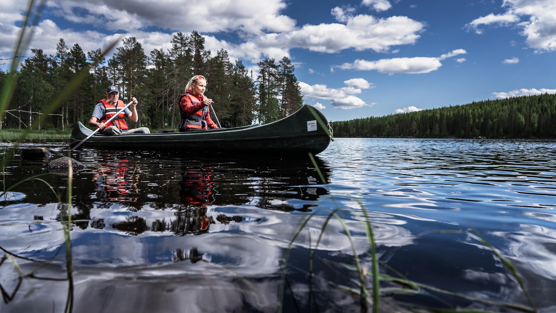 Zwei Personen paddeln in einem Kanu, tief über der Wasseroberfläche fotografiert, im Hintergrund Kiefern und blauer Himmel.