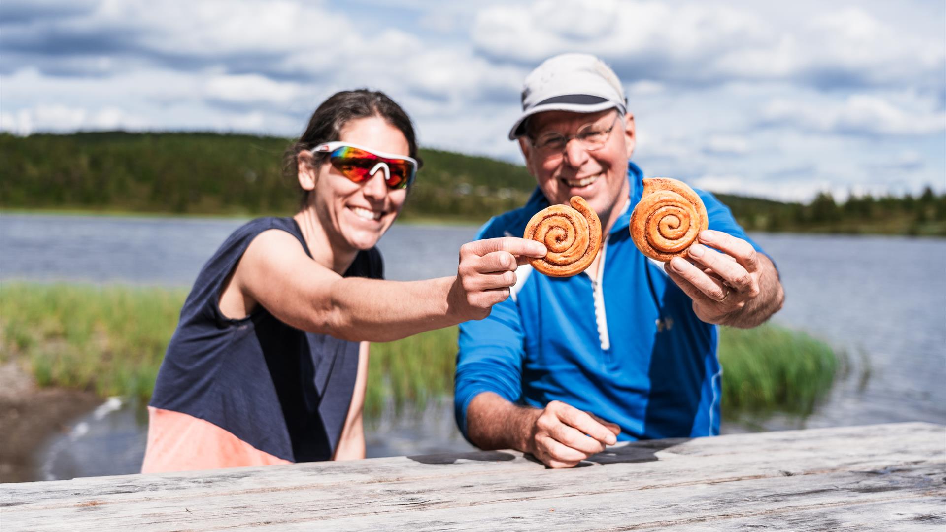 Eine Frau und ein Mann lächeln und halten zwei Zimtschnecken in die Kamera, im Hintergrund ein See.