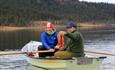 Two men in a rowboat. One steers the boat and the other shows the catch.