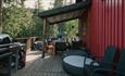 Terrasse ved en rød trebygning i skogsomgivelser, med sittegruppe og grill klart til bruk.