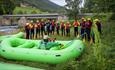 Gruppe med raftingutstyr samlet rundt en grønn flåte ved elvebredden, med bro og dal i bakgrunnen.