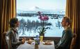 A couple eat a meal sitting in front of a window with a view of snow covered mountains