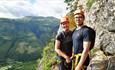 Two people in climbing gear standing by a cliff with mountain views.