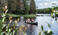 Zwei Personen paddeln in einem Kanu auf einem ruhigen Waldsee, im Vordergrund unscharfes Schilfgras.
