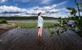 Eine Frau, in ein Handtuch gehüllt, steht im flachen Wasser eines Sees und blickt zur Seite, dahinter Schilf und Wald.