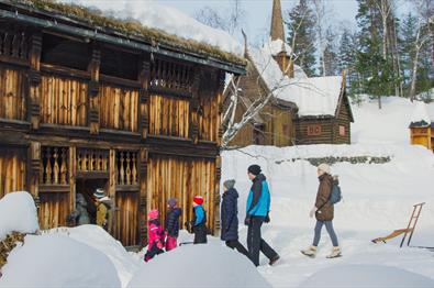 Voksne og barn går inn i tømmerhus ved Garmo stavkirke.