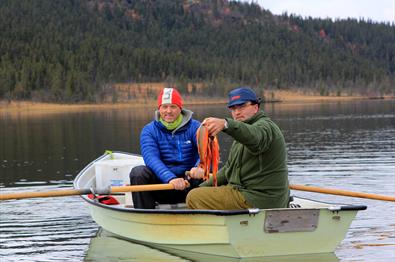 Two men in a rowboat. One steers the boat and the other shows the catch.