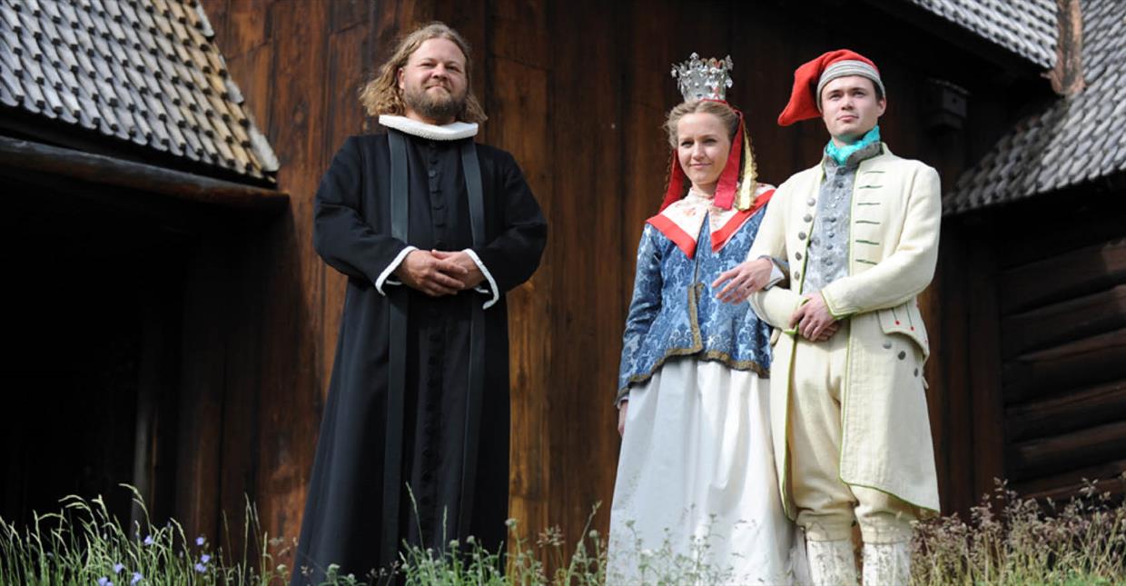 Prest og brudepar utenfor Garmo stavkirke på Maihaugen Museum, iført tradisjonelle klær i henhold til tidsperioden.