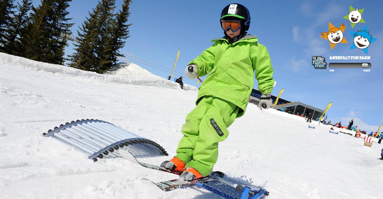 Liten gutt i grønne klær står på ski nedover railsene i barnebakken.