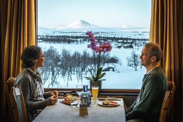 A couple eat a meal in front of a window with a view of snowy mountains
