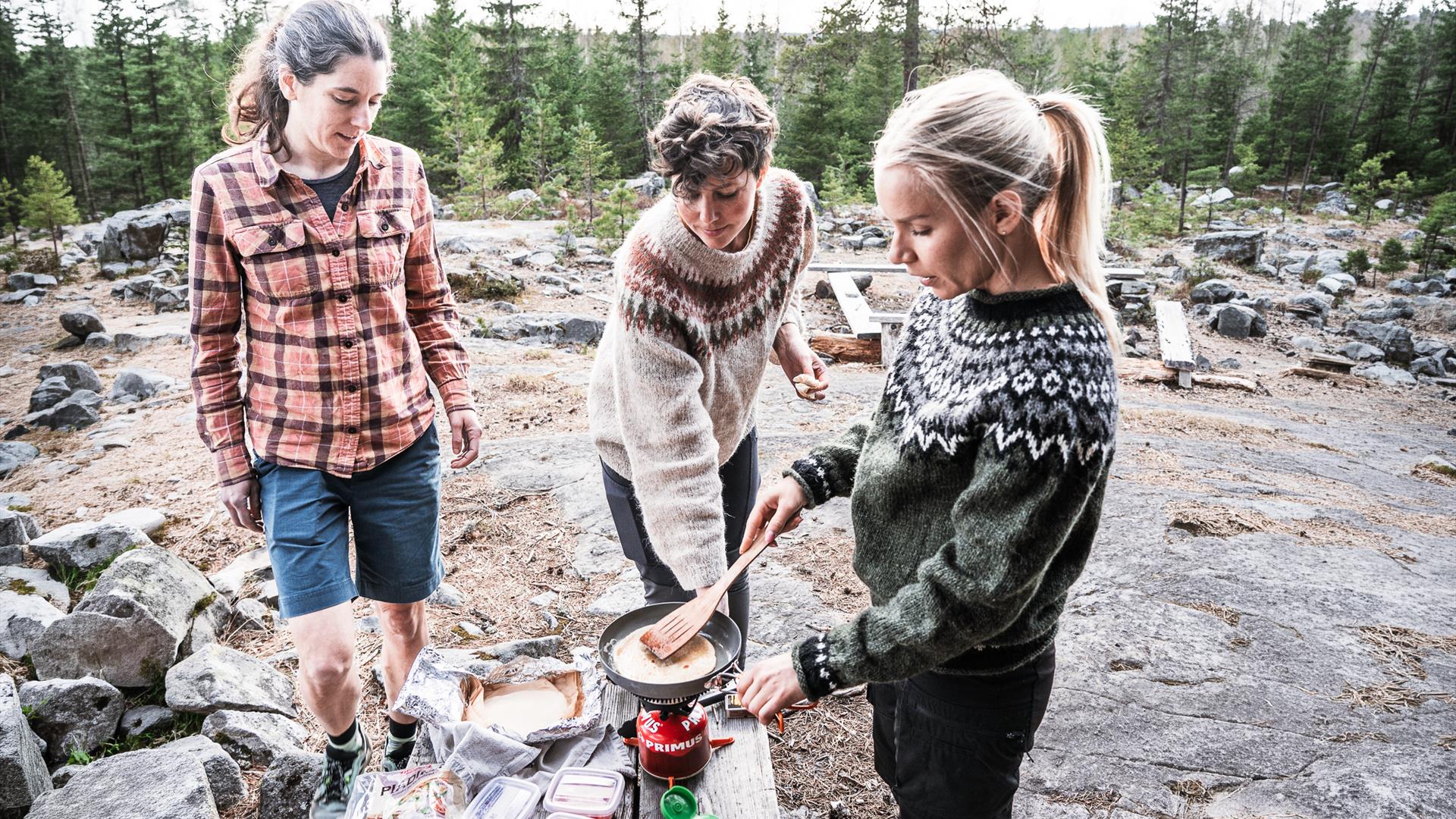 Tre personer lager mat utendørs ved bålplass. Tre personer lager mat utendørs ved bålplass.