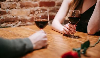 Two people sat on a wooden table, with two glasses on red wine.