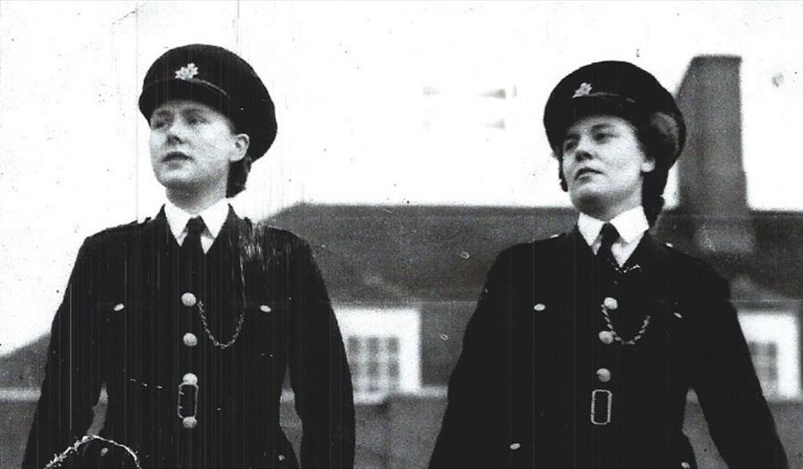 Two policewomen striding in lockstep towards the camera, in front of a large house. Black and white photograph.