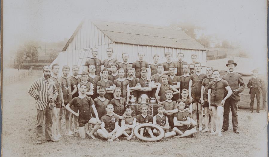 Sepia toned group photograph of Maidstone Swimming Club members, all males in swimming costumes, standing, kneeling or sitting in five rows, a life-fl