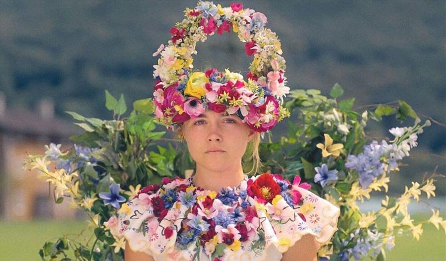 An image from the movie of Florence Pugh, a blonde, short woman, wearing a floral dress and head piece. In the background there are gardens,