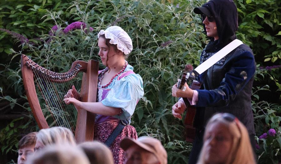 A pair of performers stand playing musical instruments in an outdoor garden environment.