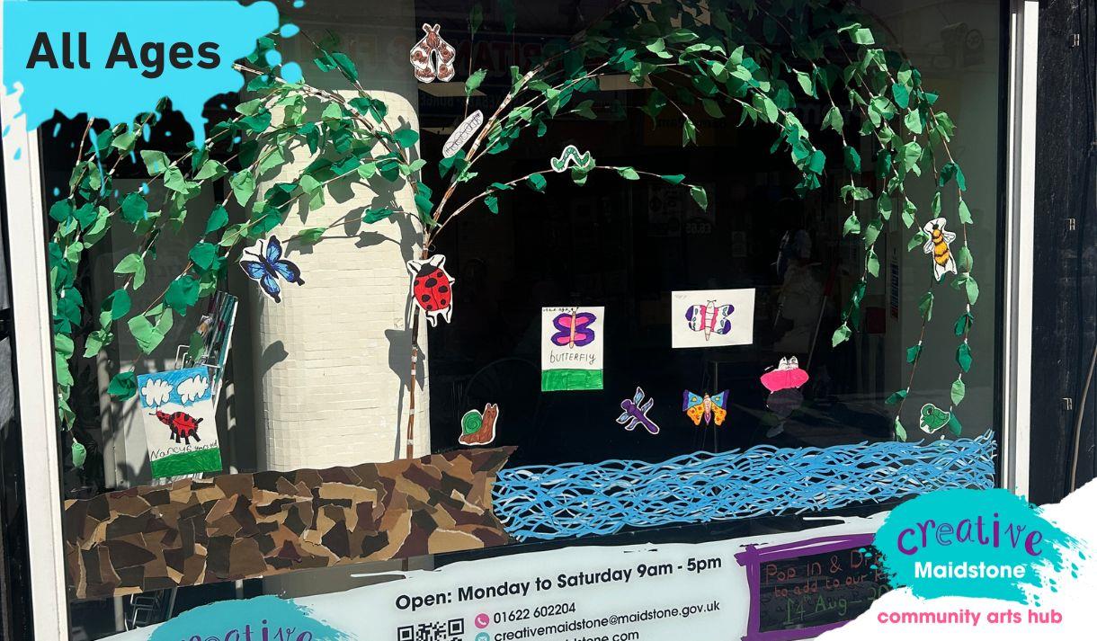 A shop window decorated with hand drawn bugs and a river with a paper willow tree