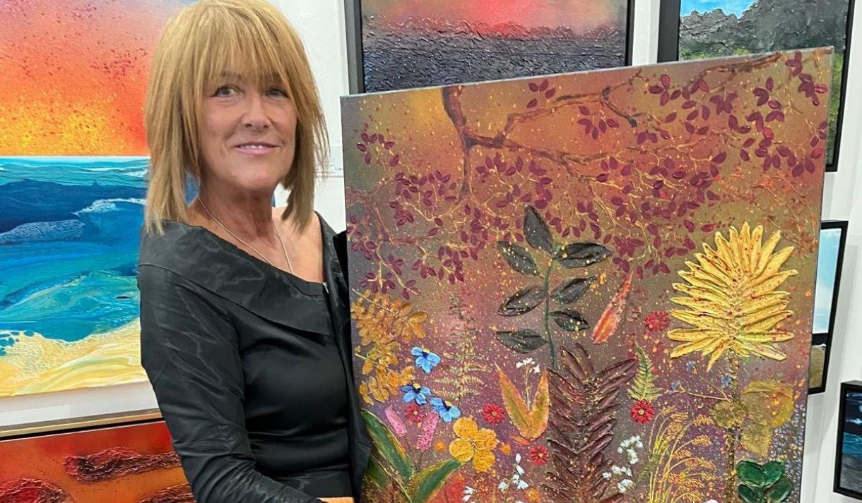 Artist Jane Burdett holding one of her artworks - an imagined landscape with a variety of plants and flowers