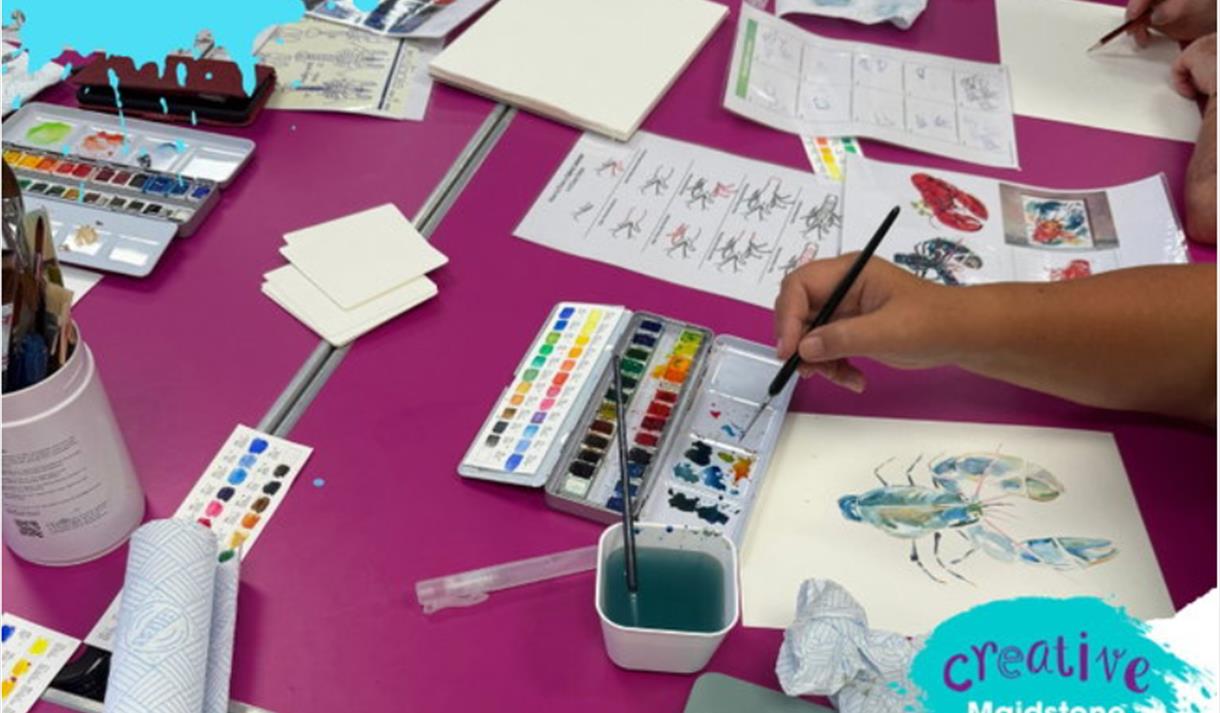 A picture of a pink table covered in paints and paint brushed and paintings of lobsters