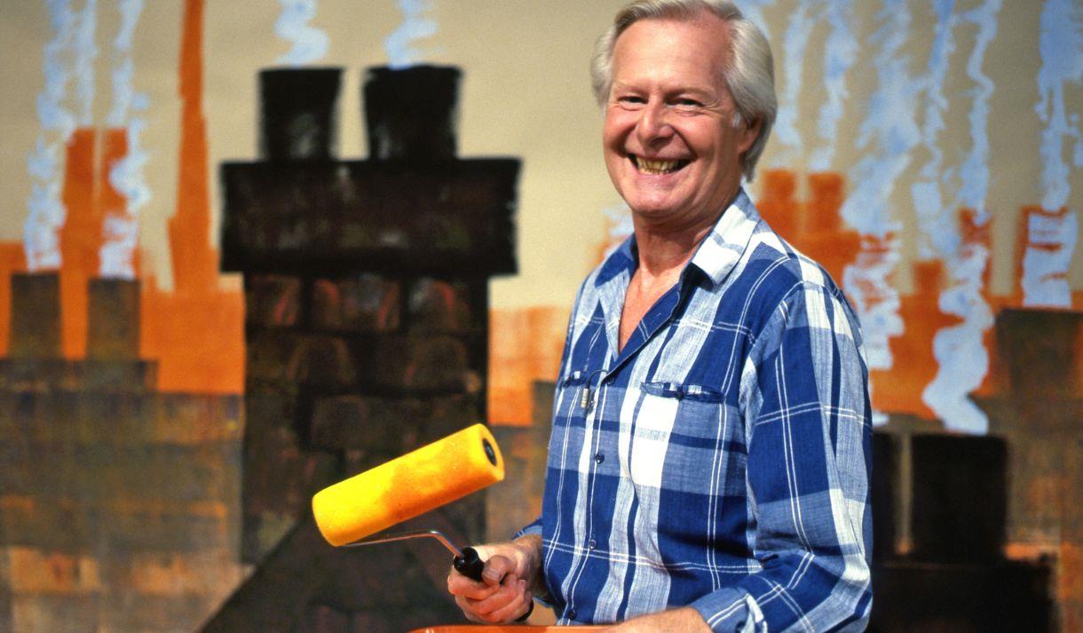 Tony Hart holding a paint tray and roller in front of a painted background of rooftops and chimneys