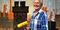 Tony Hart holding a paint tray and roller in front of a painted background of rooftops and chimneys