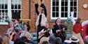 A group of performers sing and dance against the backdrop of a brick building.