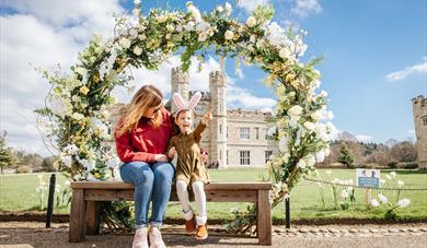 Easter Fun at Leeds Castle - Lady sitting under a flower hoop with child wearing bunny ears.