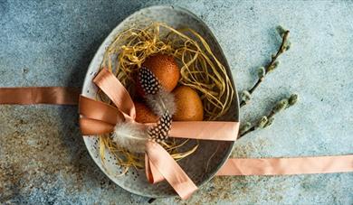 Speckled brown eggs in a bowl with straw with pussy willow and  ribbon.