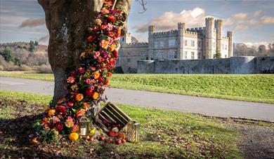 Floral arrangement in a tree with a face in the bark and lamps, apples and pumpkins on the ground. Background of Leeds Castle.
