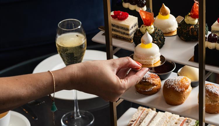 Person reaching for cake from an afternoon tea platter with champagne